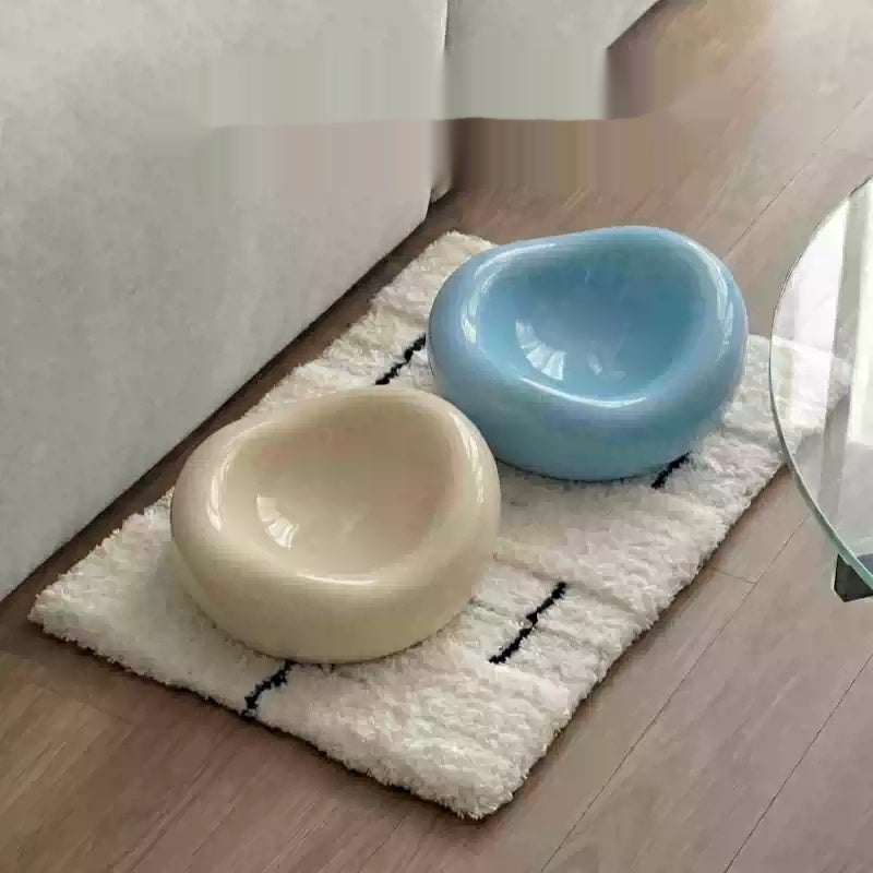 Two glossy, organic-shaped pet bowls, one cream and one light blue, placed on a white textured rug on a wooden floor next to a sofa.