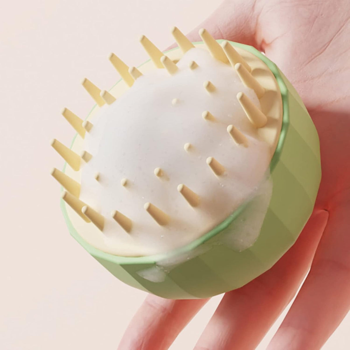 Green scalp massager held in a hand against a light pink background