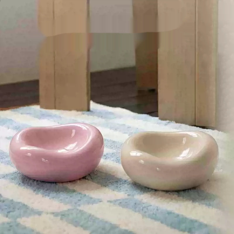 Two small glossy ceramic dish holders in pastel pink and cream, placed on a blue-and-white striped rug with wooden furniture legs in the background.