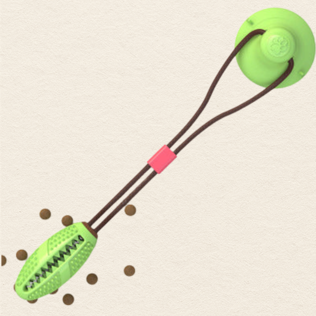 A green dog chew toy with a suction cup on one end, brown elastic cords, and a small red connector, shown on a light beige textured background with a few scattered brown chew pieces.