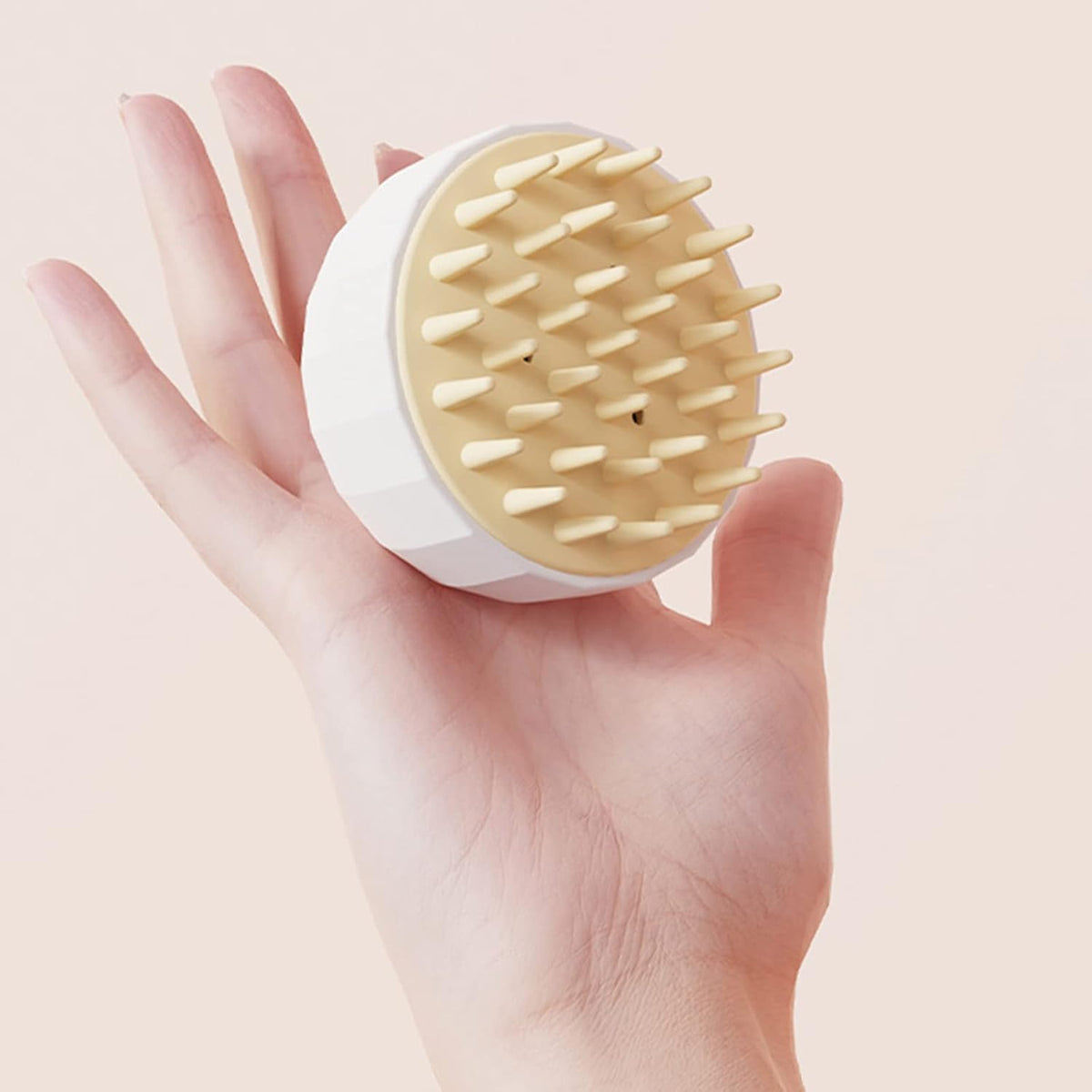 Hand holding a round, white and beige scalp massager against a light pink background